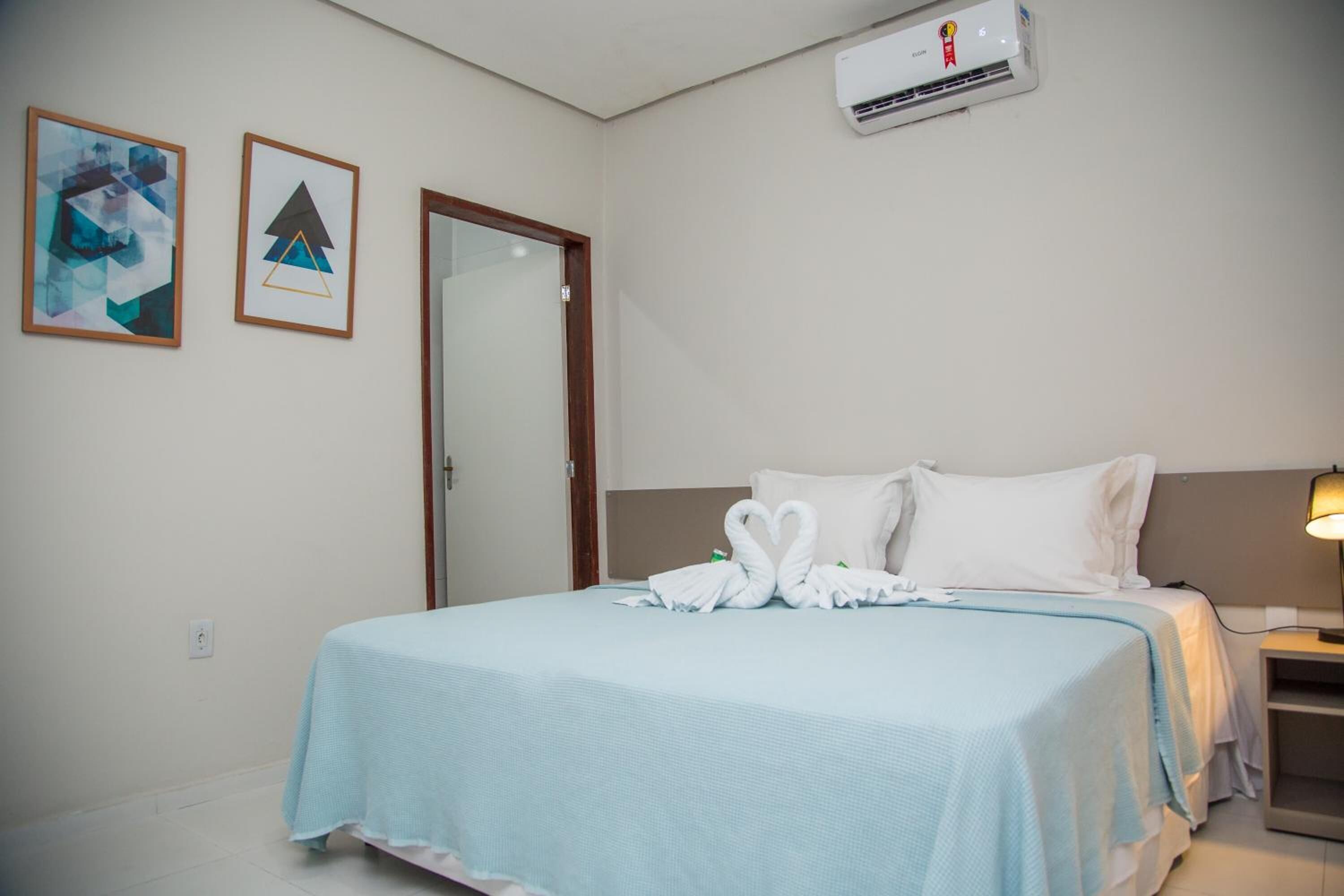 A hotel room with a bed, a nightstand, a lamp, an air conditioner, and two framed pictures on the wall.