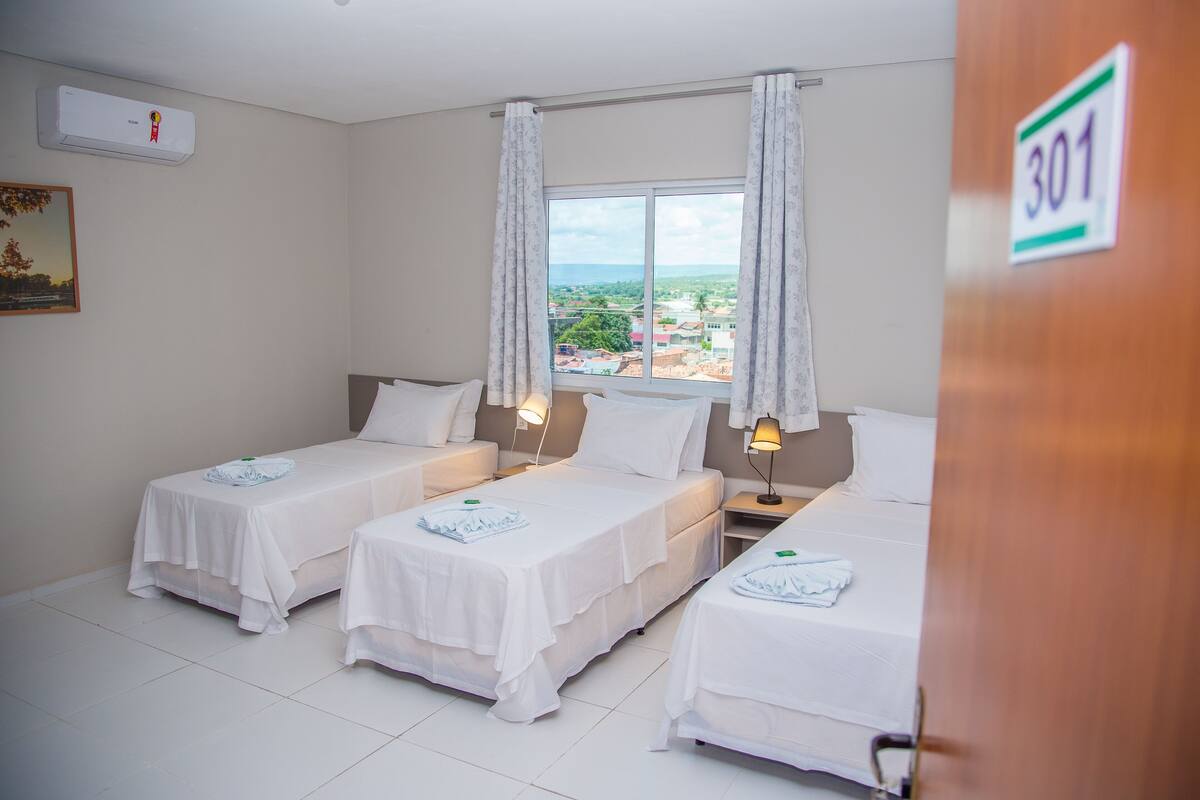 Habitación de hotel con tres camas individuales, una ventana con cortinas y vistas a la ciudad, además de un cartel con un número en la puerta.