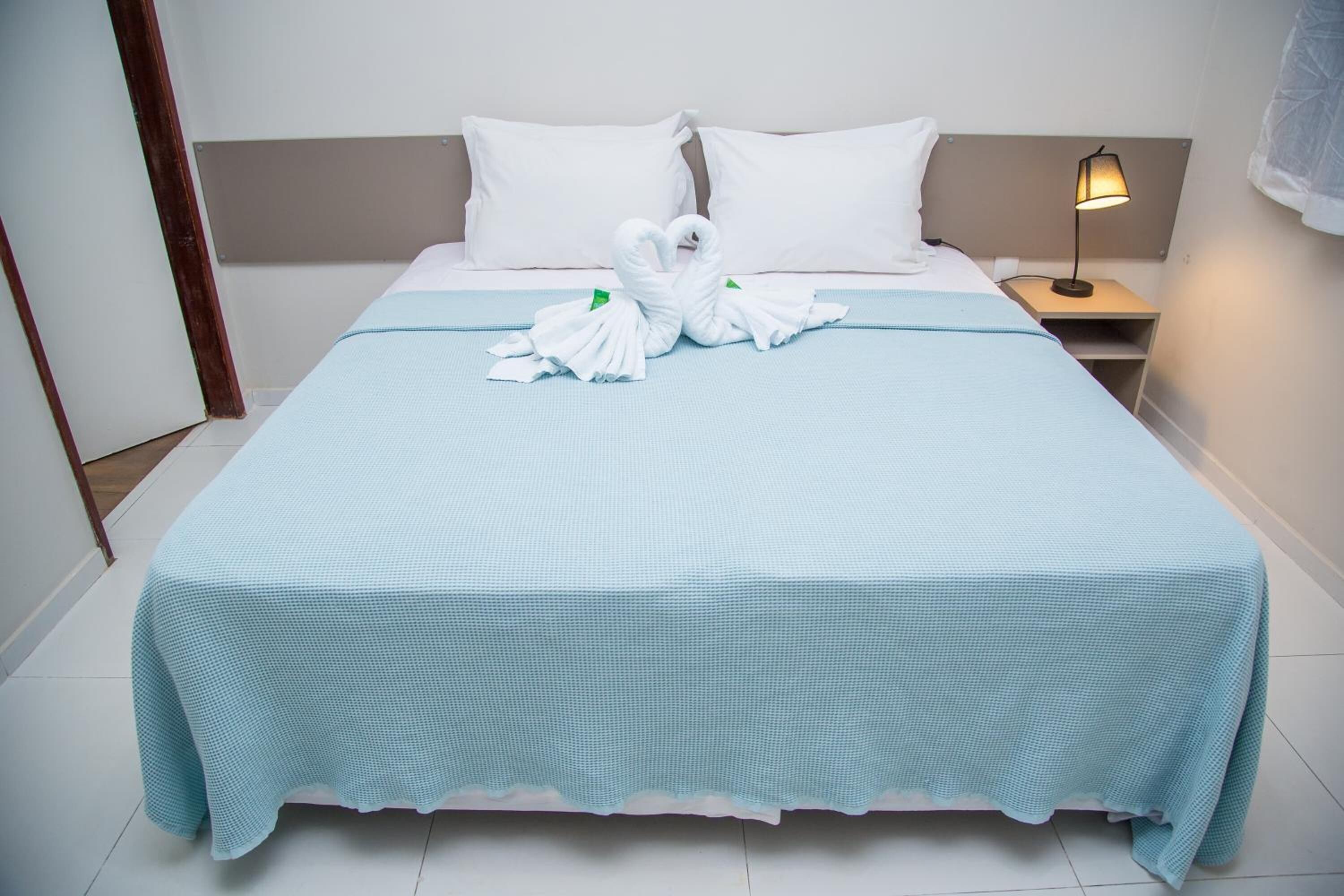 Una cama bien tendida con una colcha azul claro, dos almohadas blancas y un par de toallas blancas dispuestas en forma de corazón sobre la cama.