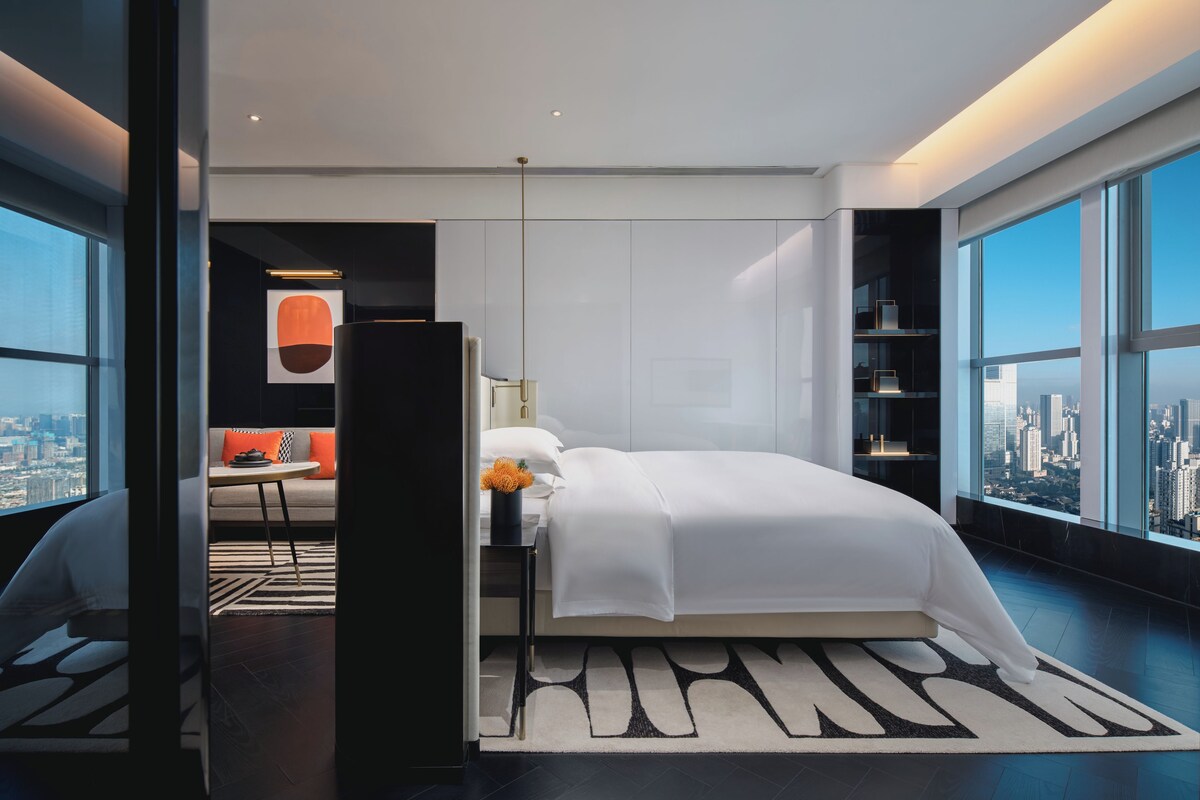 Een moderne hotelkamer met een groot bed, een zithoek met een bank en een tafel, en een stadsgezicht door grote ramen.