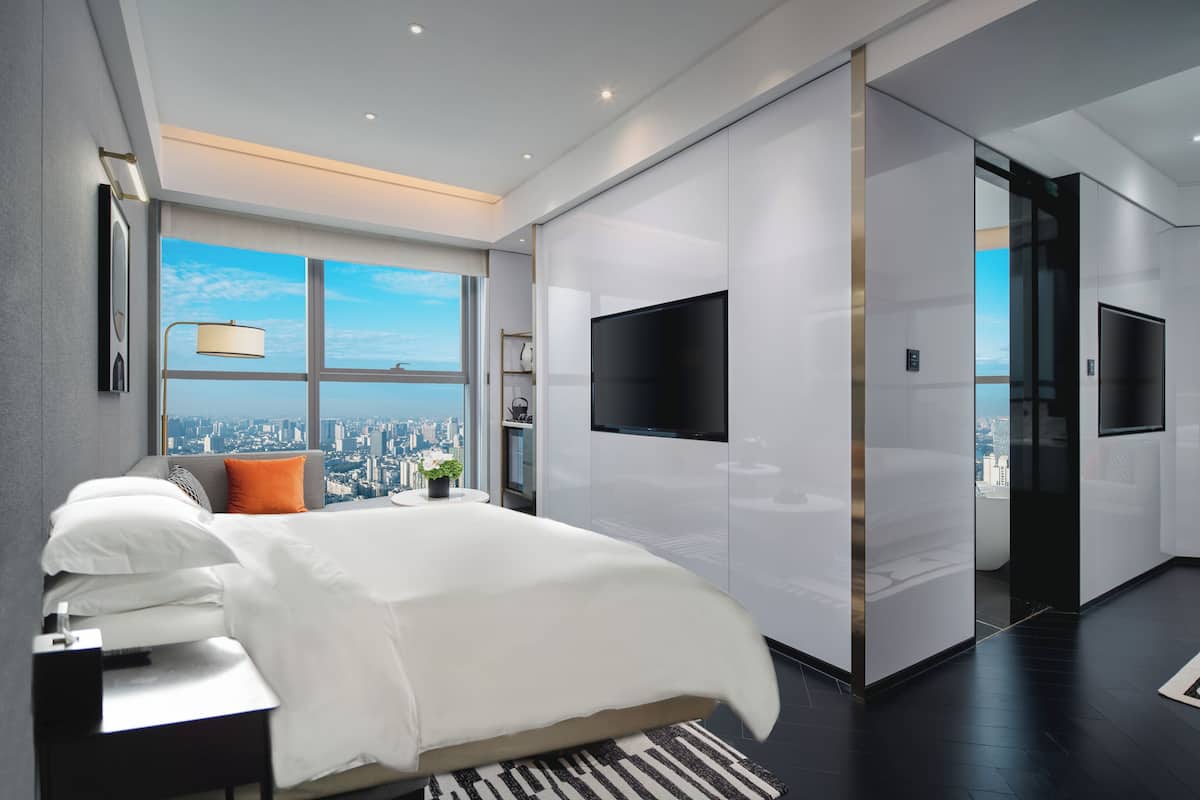 Une chambre d’hôtel moderne dotée d’un grand lit, d’un téléviseur à écran plat, d’une vue sur la ville et d’une porte coulissante donnant accès à la salle de bain.