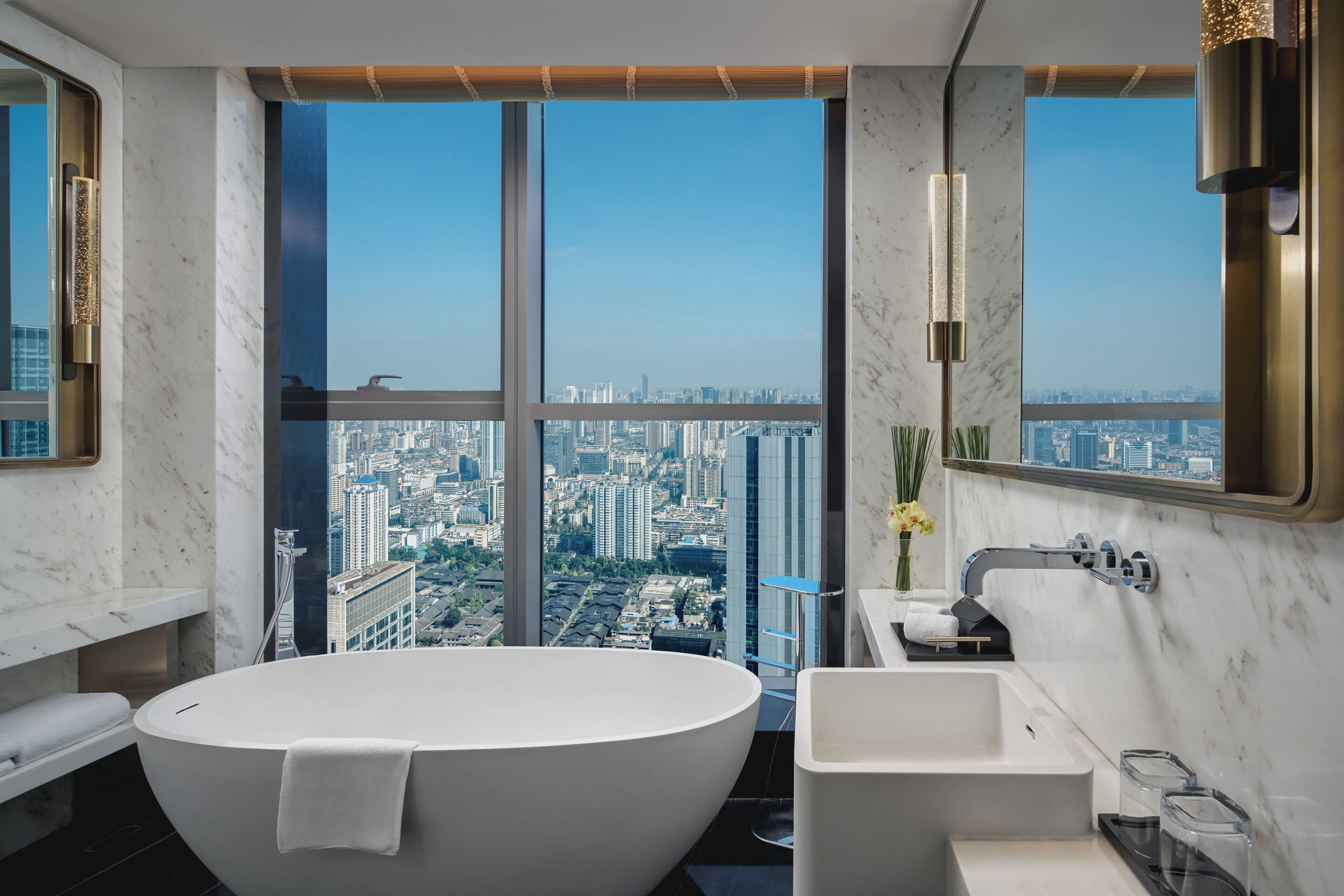 A modern bathroom with a freestanding bathtub, a sink, and a large window offering a city view.