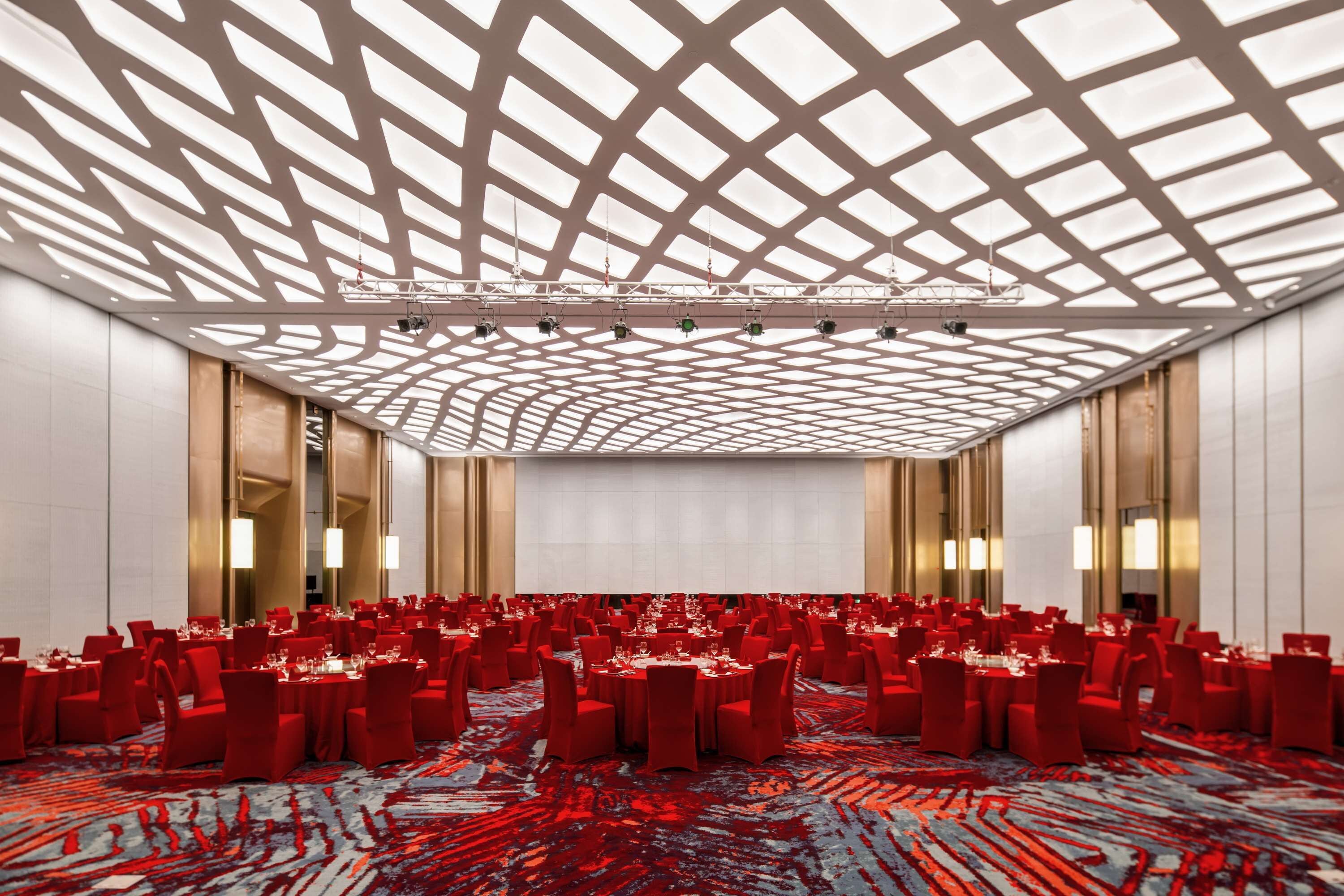 Ein großer Konferenzraum mit hoher Decke und einem geometrischen Muster. Tische sind mit roten Stühlen und weißen Tischdecken gedeckt.