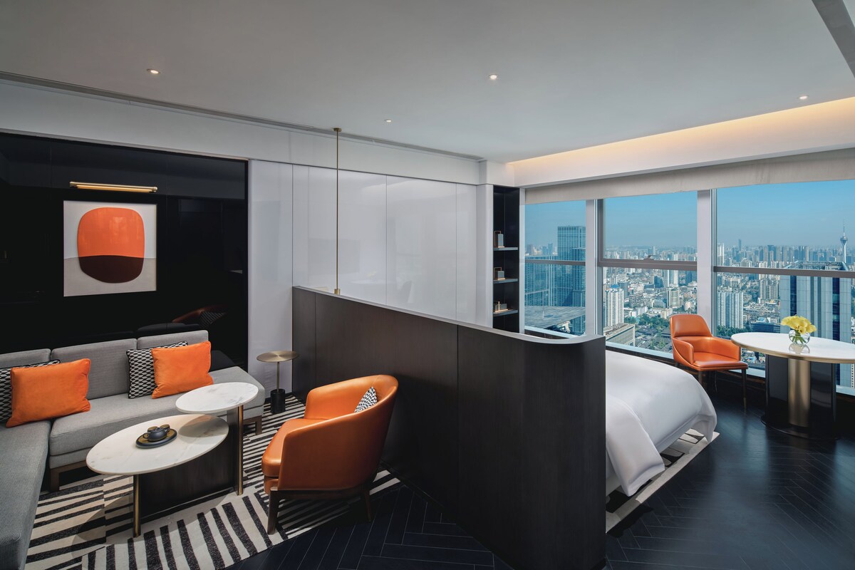 Een moderne hotelkamer met een hoekbank, een ronde salontafel, een oranje fauteuil, een wit bed en een stadszicht door grote ramen.