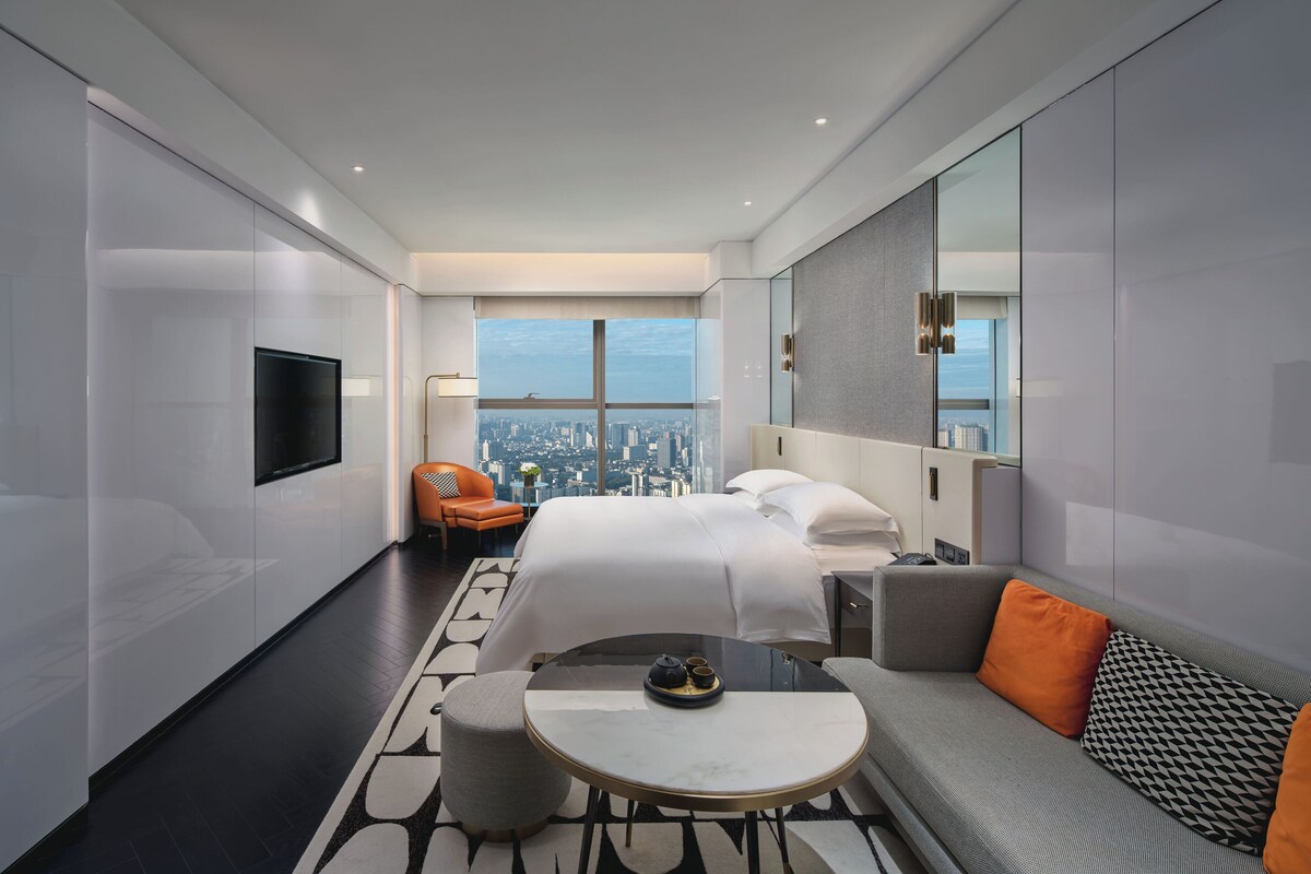 Een moderne hotelkamer met een groot bed, een bank, een klein rond tafeltje en uitzicht over de stad.