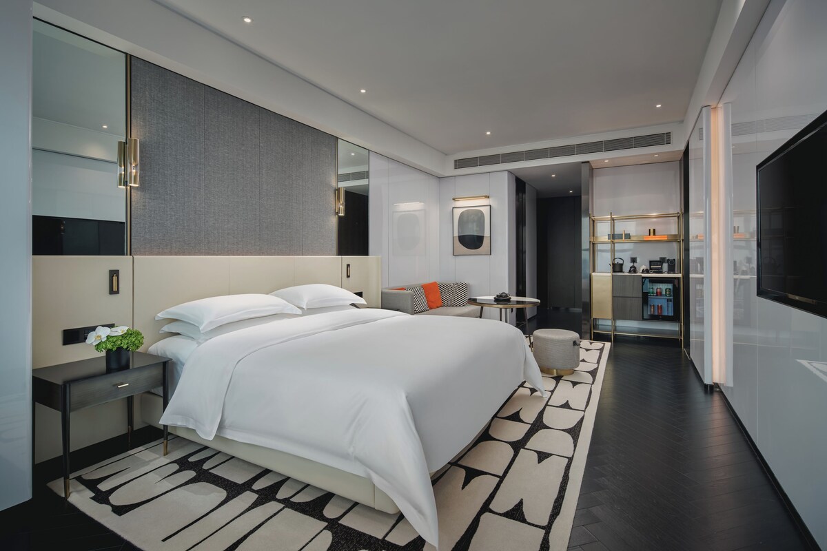 Een moderne hotelkamer met een groot bed, een nachtkastje, een klein bureau, een stoel, een bank, een televisie en een badkamer zichtbaar door een glazen deur.