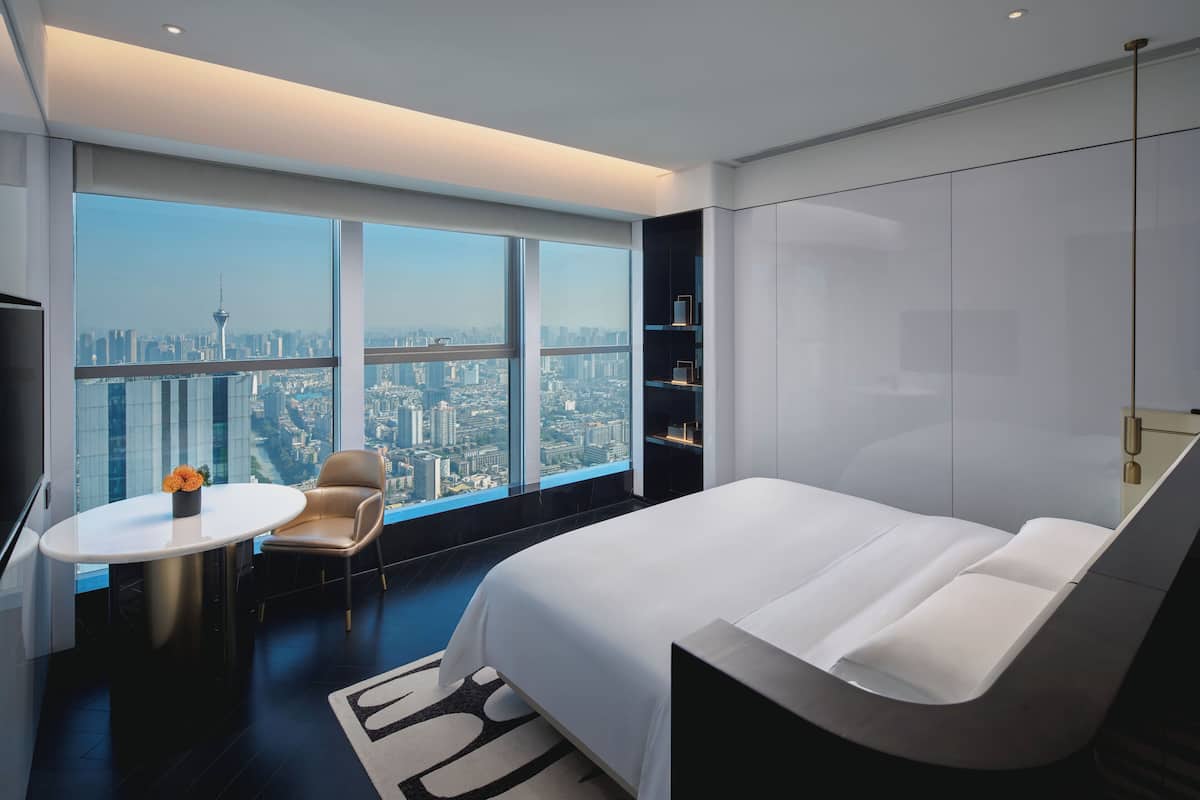 Une chambre d’hôtel moderne avec un grand lit, une table ronde, une chaise et une vue sur le paysage urbain, visible à travers de grandes fenêtres.