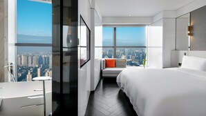 Una habitación de hotel moderna con una cama grande, una zona de estar con vistas y un paisaje urbano visible a través de la ventana.