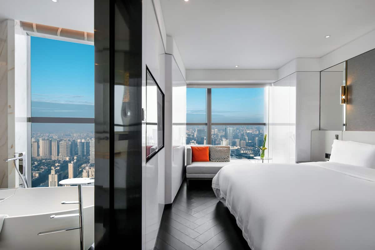 Une chambre d’hôtel moderne dotée d’un grand lit, d’un coin salon avec vue et d’une fenêtre donnant sur le paysage urbain.