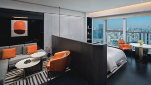 Habitación de hotel moderna con sofá modular, una mesita redonda, un sillón naranja, una cama blanca y vistas a la ciudad a través de amplias ventanas.