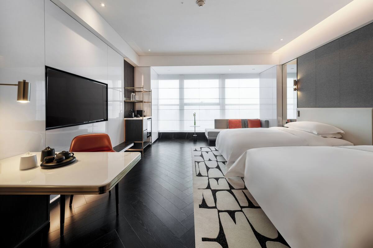 Une chambre d’hôtel moderne équipée d’une télévision à écran plat, d’un bureau avec une chaise, de deux lits, d’un canapé et d’une grande fenêtre laissant entrer la lumière naturelle.