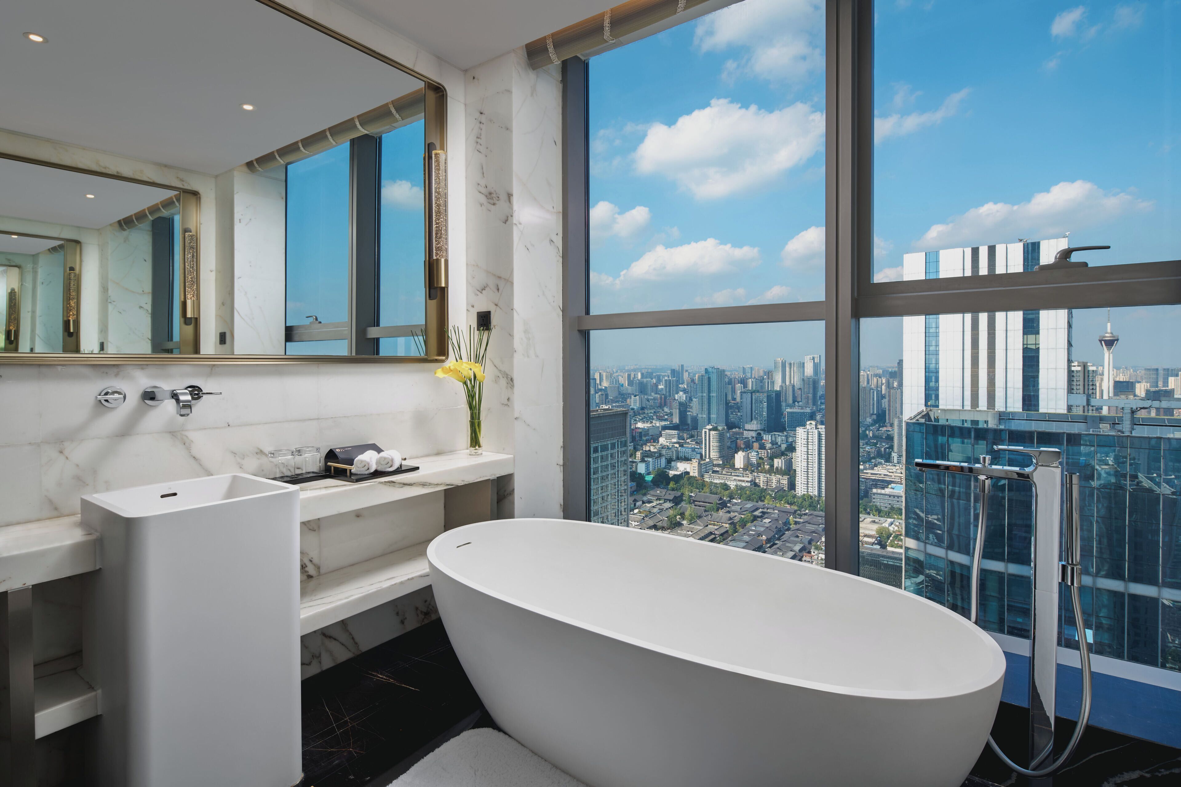 Un baño moderno con una bañera independiente, un espejo grande y vistas a la ciudad a través de amplias ventanas.