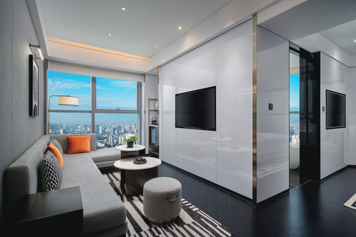 Een moderne woonkamer met een bank, poef en salontafel, een flatscreen-televisie aan de muur en een groot raam met uitzicht op de stad.