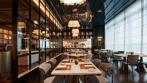 Interior moderno de restaurante con techos altos, ventanas amplias y una zona de comedor central con múltiples mesas y sillas.