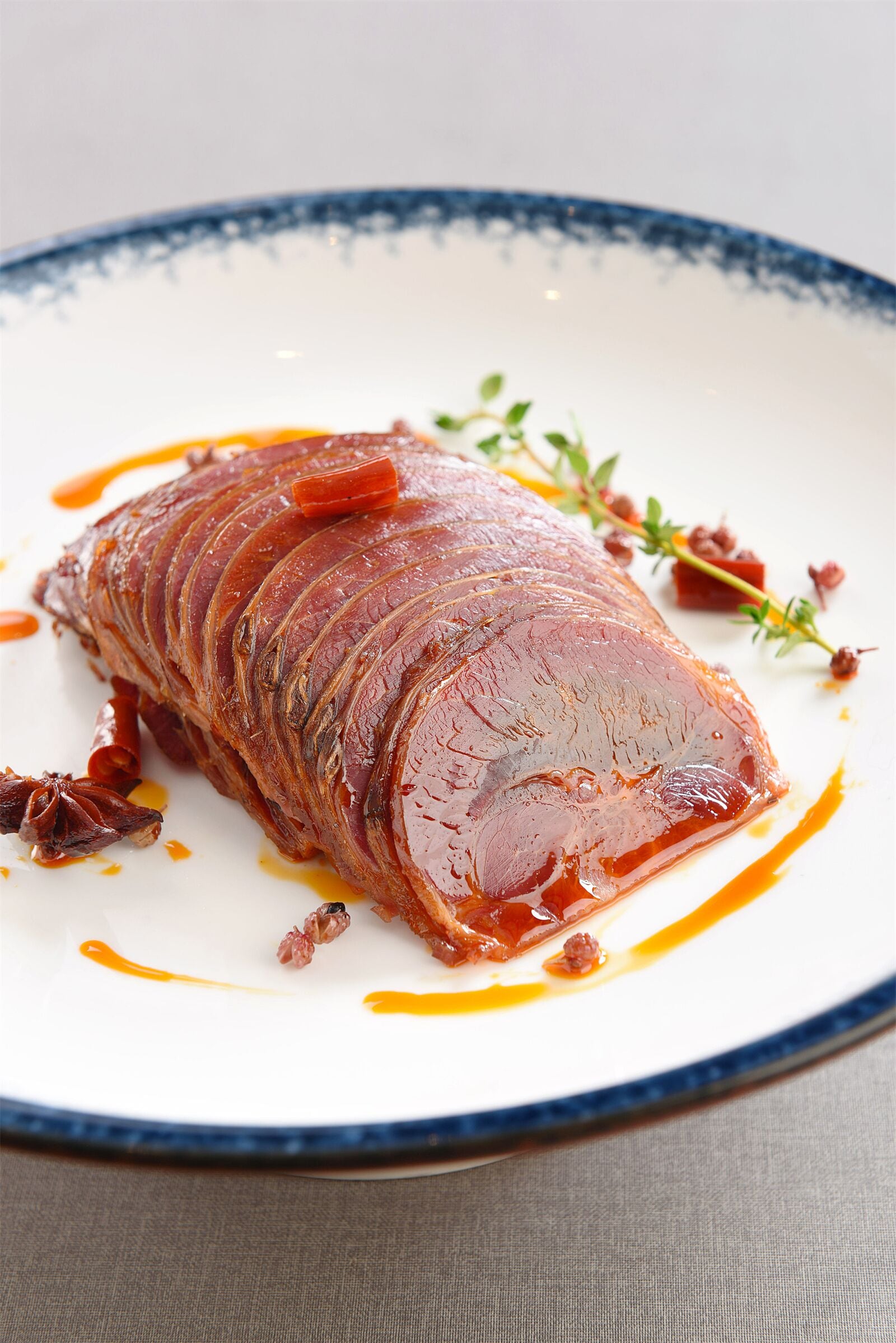 Un jamón grande y brillante sobre un plato blanco con borde azul, adornado con hierbas y salsa.