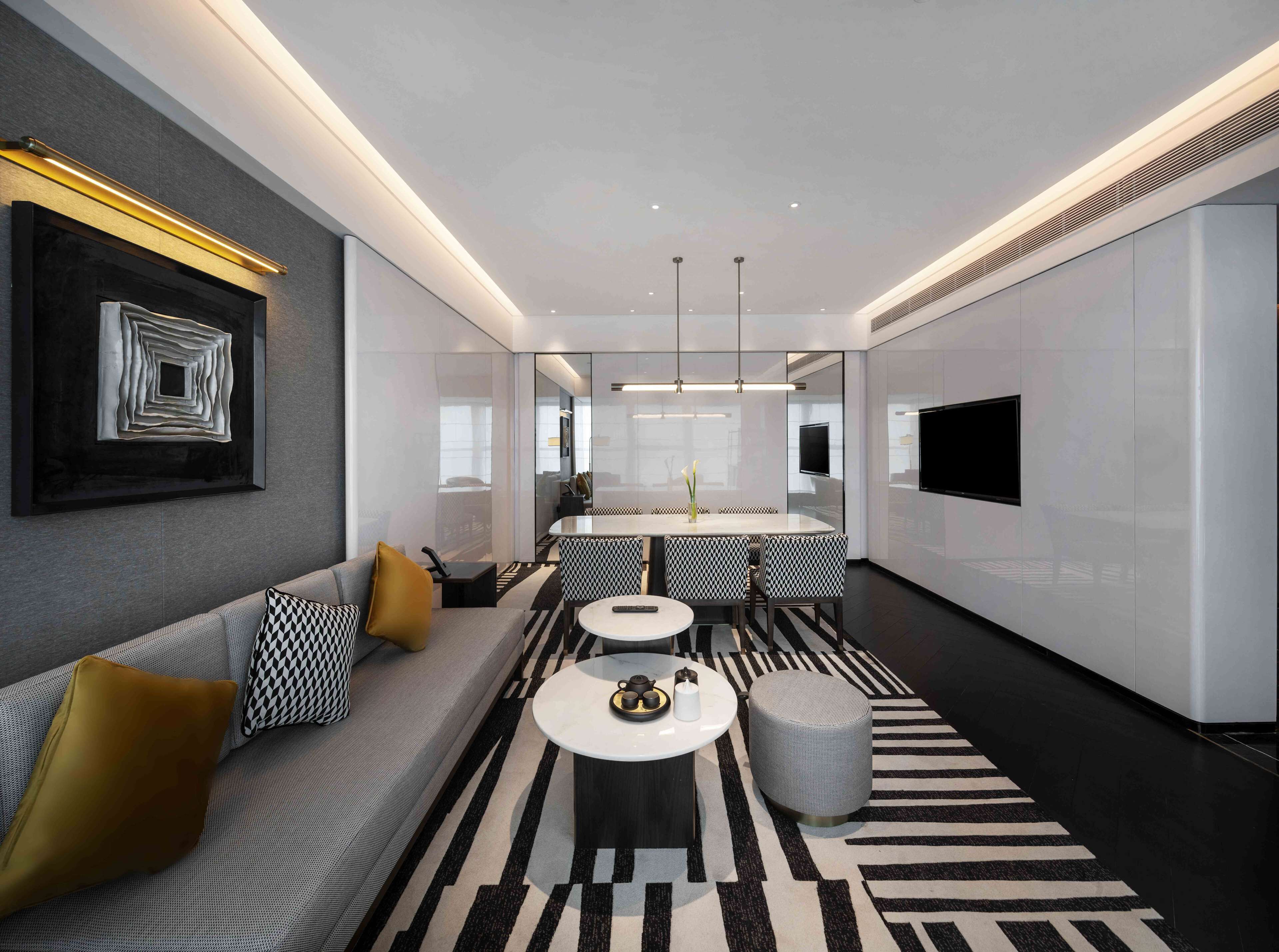A modern hotel room with a sofa, a round coffee table, a patterned rug, and a TV mounted on the wall.