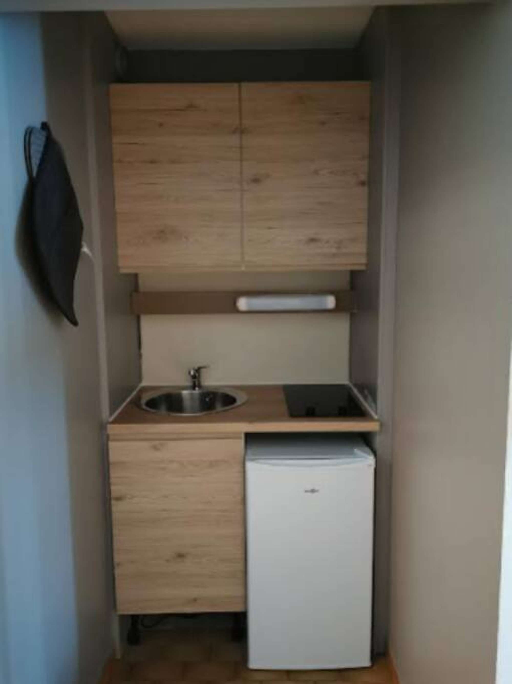 Une cuisine compacte avec des meubles de rangement en bois, un évier, un petit réfrigérateur et un objet noir suspendu.