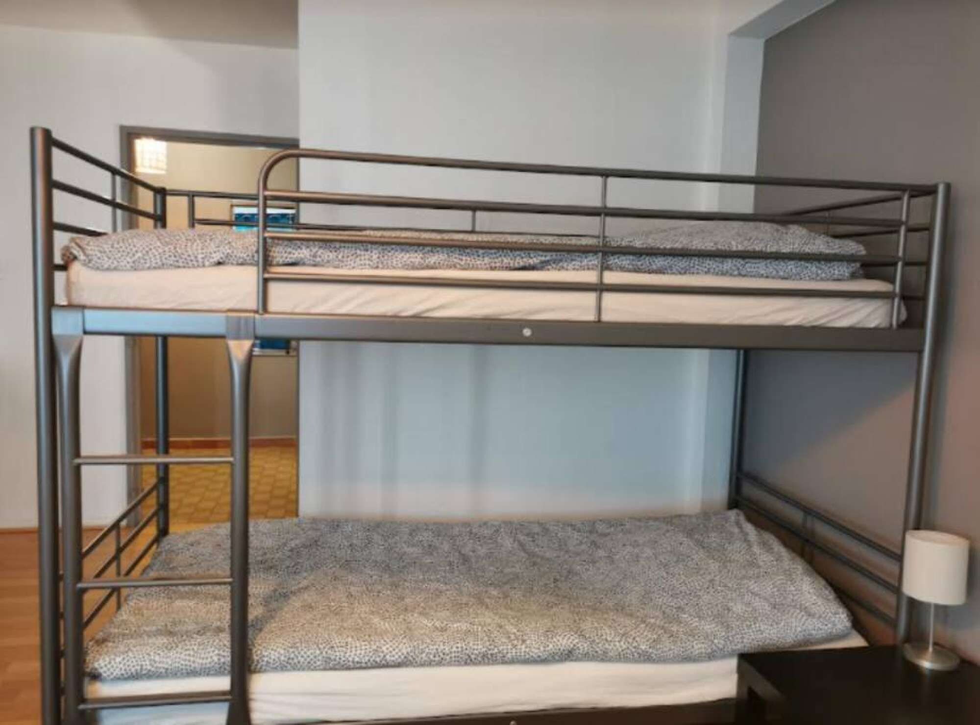 A bunk bed with two levels, a small table with a lamp, and a plain wall.