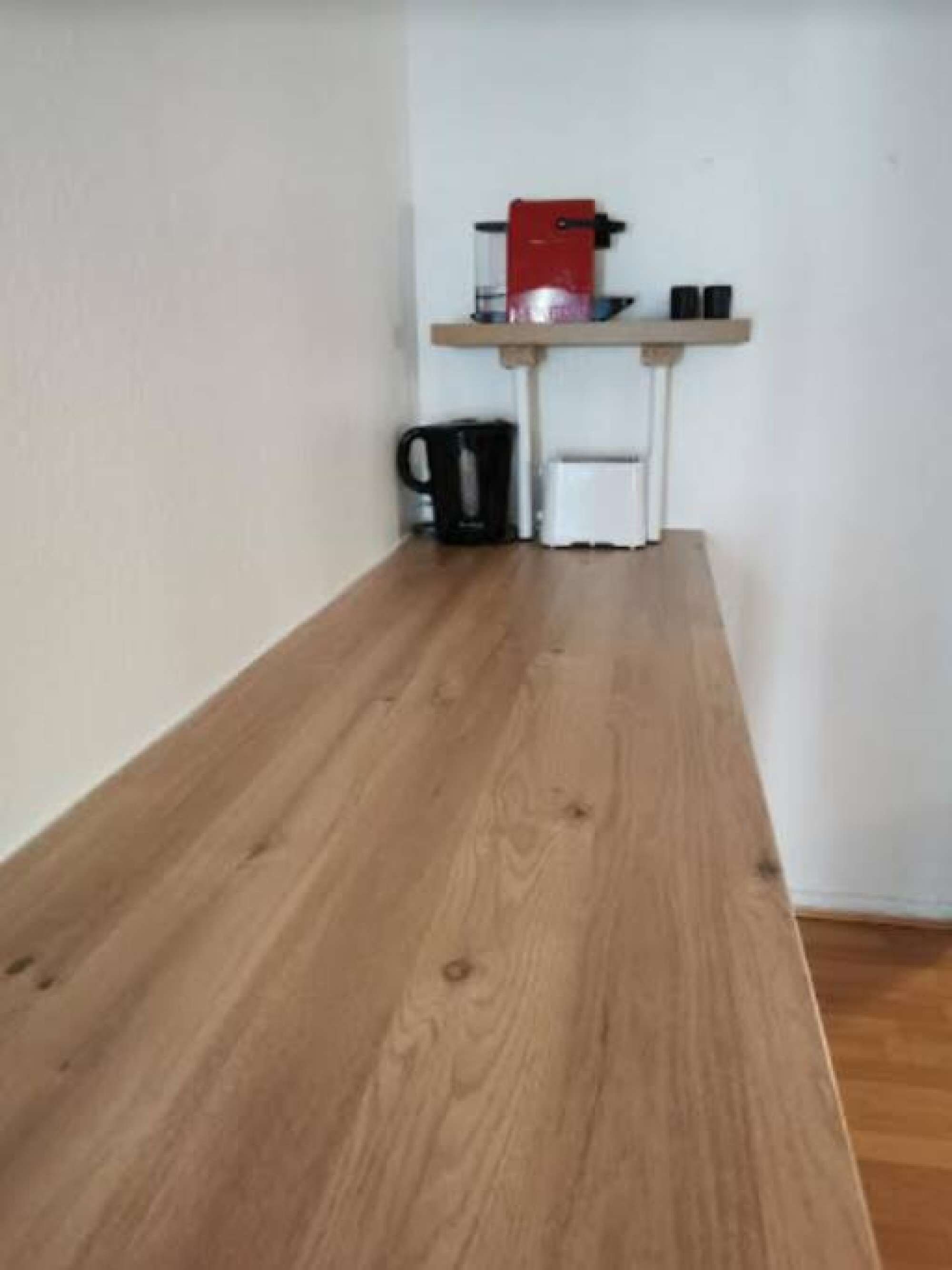Une table en bois dans une pièce dont le mur est blanc, avec une petite étagère sur laquelle se trouvent un appareil rouge et un objet noir.