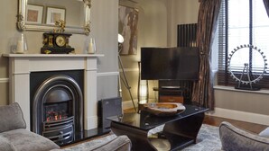 Una sala de estar con chimenea, un televisor sobre un mueble bajo, un espejo grande y un ventanal con cortinas.