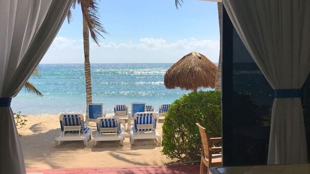 Vista de la playa desde una habitación con un gran ventanal, que incluye un paraguas de paja, varias sillas de descanso y vista al océano.