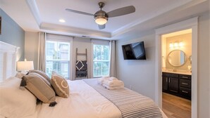 Un dormitorio con cama, ventilador de techo, televisor montado en la pared y baño visible a través de una puerta abierta.