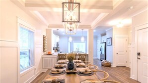 Un comedor con una mesa de madera dispuesta para seis personas, rodeada de sillas, una araña y vistas a la cocina.
