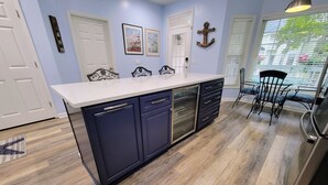 Cocina con isla central, armarios azul oscuro, lavavajillas y refrigerador. Hay dos puertas, una ventana con persianas y una silla.