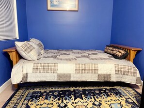 Une chambre avec un mur bleu, un lit recouvert d’une courtepointe, deux oreillers, une table de chevet en bois et un tapis à motifs.