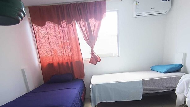 Ein Zimmer mit zwei Betten, einem Fenster mit roten Vorhängen, einer Klimaanlage und einem grünen Stuhl.