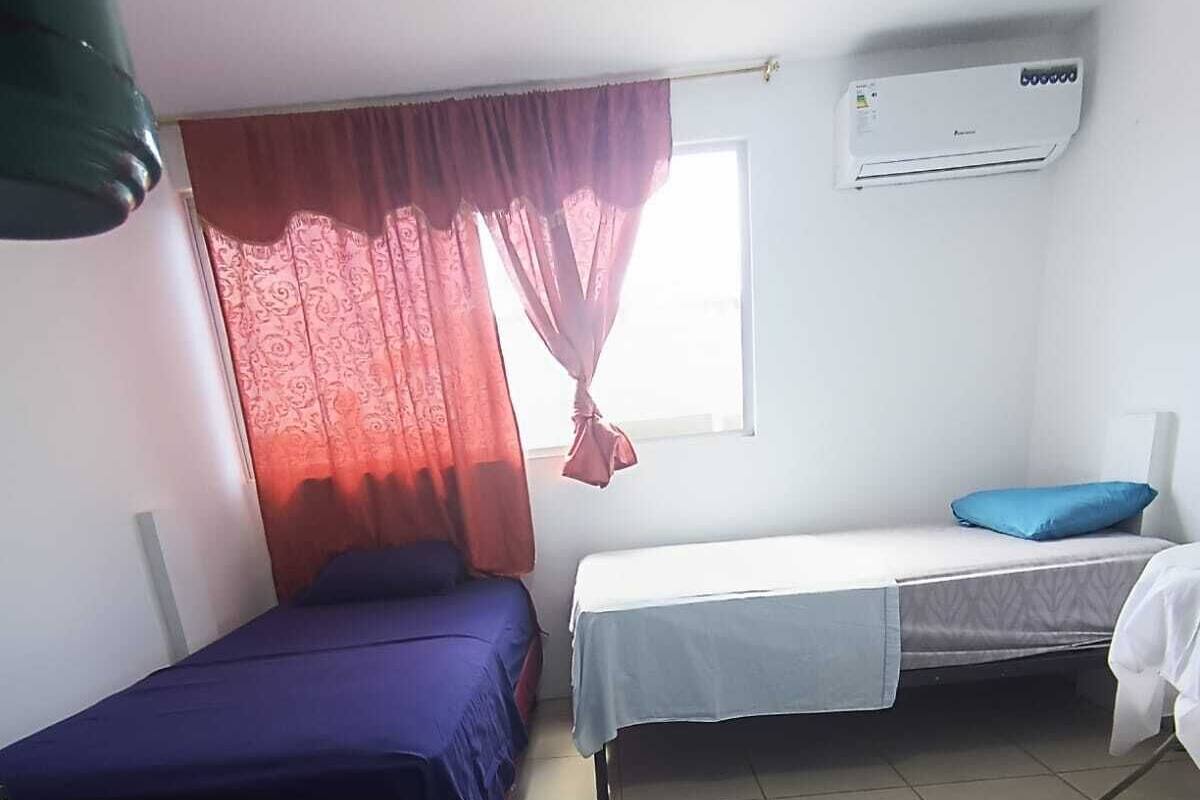 Une chambre avec deux lits, une fenêtre avec des rideaux rouges, un climatiseur et un fauteuil vert.