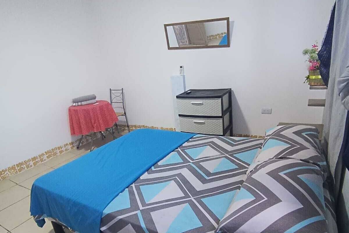 Une chambre à coucher avec un lit, une petite table recouverte d’une nappe rouge, une commode et un miroir accroché au mur.
