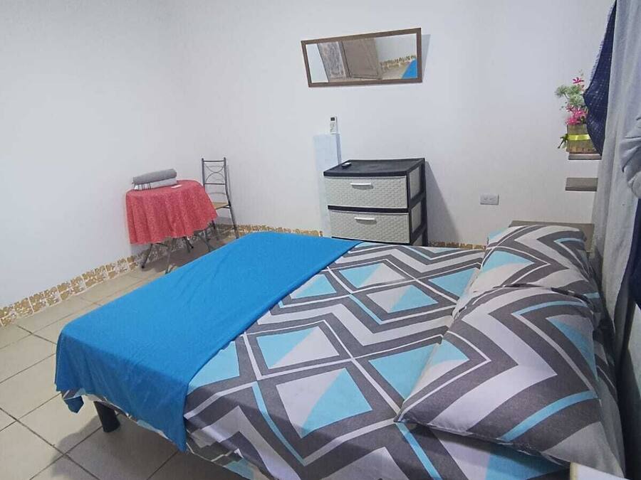 Une chambre à coucher avec un lit, une petite table recouverte d’une nappe rouge, une commode et un miroir accroché au mur.
