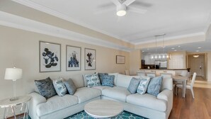 Un salon moderne avec un canapé d’angle, une table basse ronde et un tapis à motifs. On y trouve également un coin repas avec une table et des chaises, et une cuisine à l’arrière-plan.