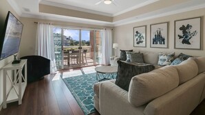 Un salon avec un canapé beige, un téléviseur, un ventilateur de plafond et une vue sur un balcon équipé d’une table et de chaises.