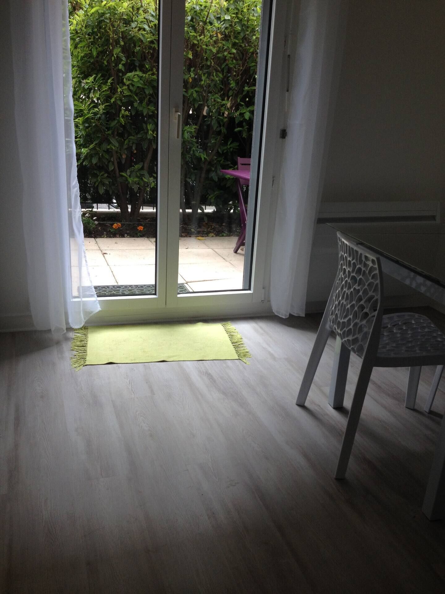 Une pièce dotée d’une porte-fenêtre coulissante qui donne sur un patio. On y trouve un tapis vert près de la porte, et une chaise est placée sur la droite.