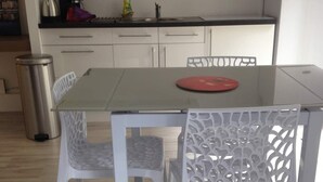 Una cocina moderna con armarios blancos, un hervidor de acero inoxidable, una cafetera y un lavabo. Hay una mesa de comedor con dos sillas, una con un respaldo decorativo.