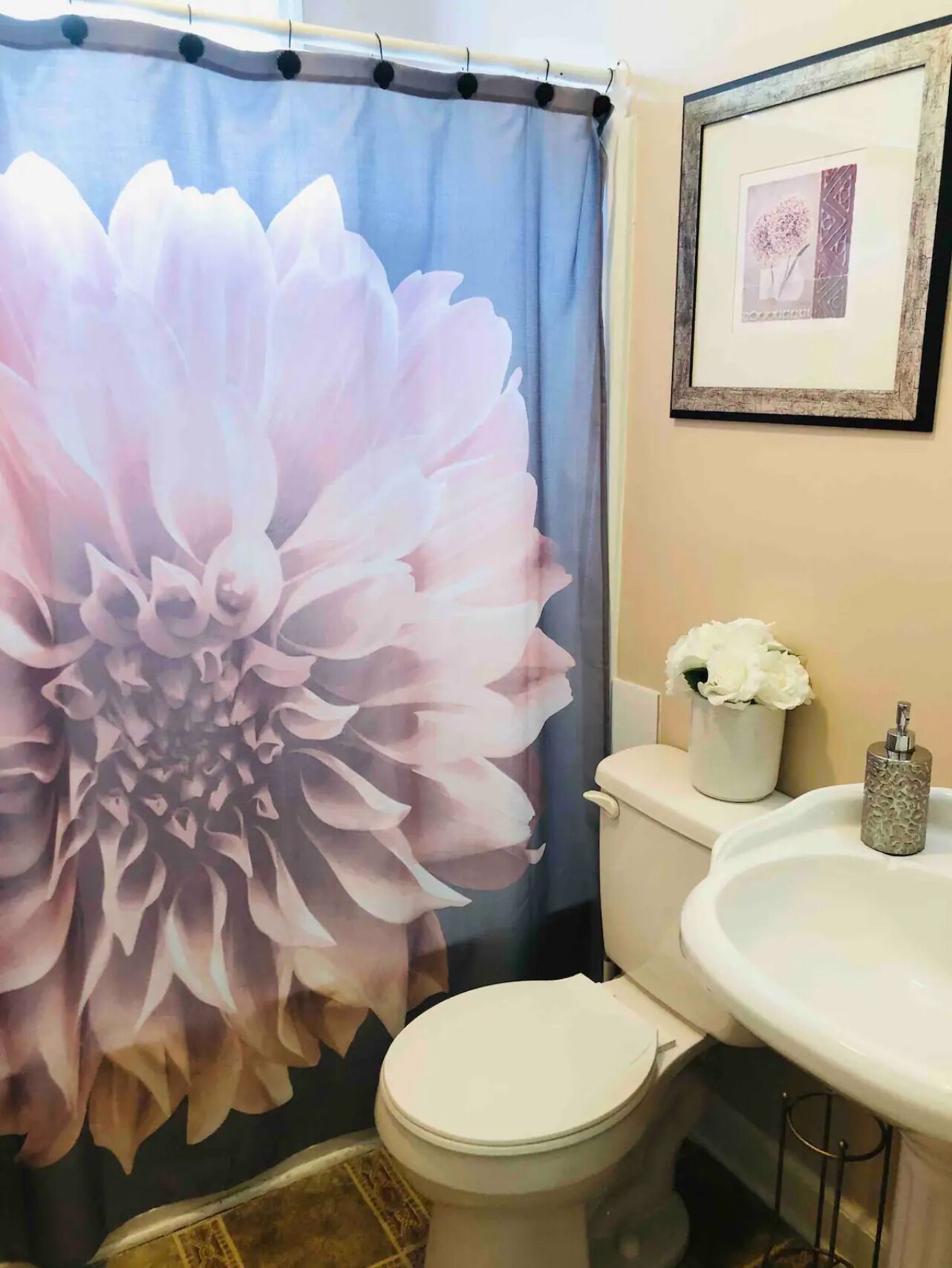 Um banheiro com cortina de chuveiro estampada com flores, vaso sanitário, pia com dosador de sabão e um quadro na parede.