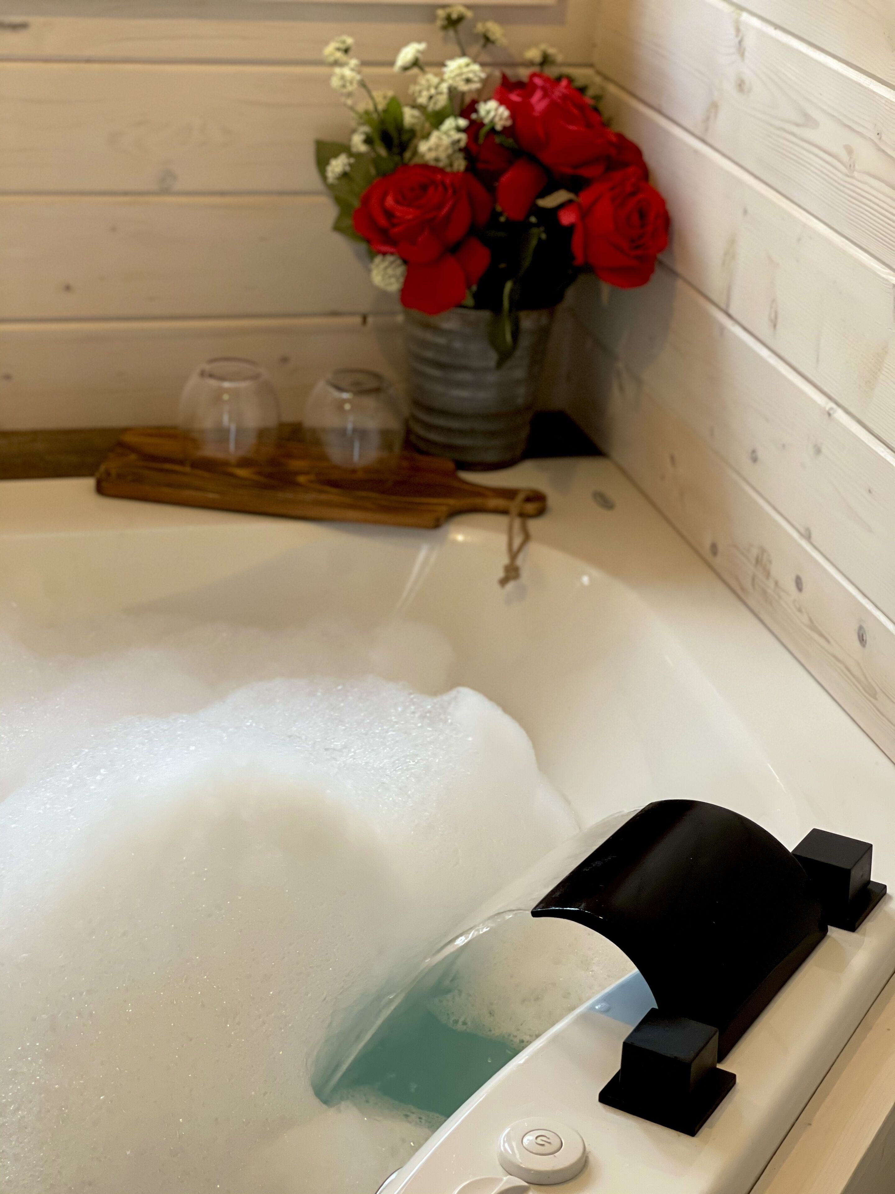 A bathtub with bubbles, a glass of water, a black object, and a vase with red roses.