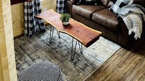 A cozy cabin interior with a brown leather sofa, checkered curtains, a wooden table with a plant, and a 'Welcome to our neck of the woods' sign.