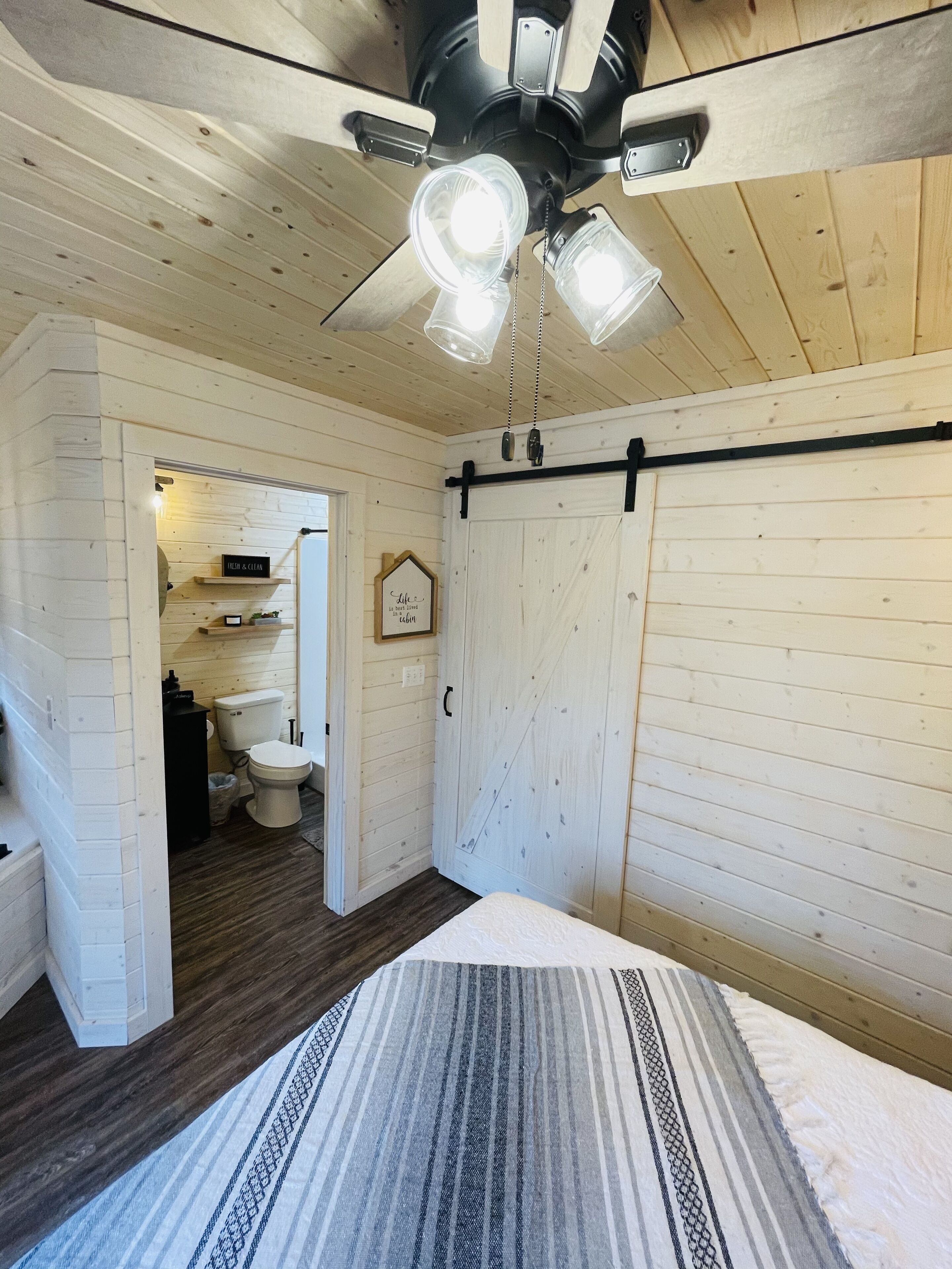 Ein Schlafzimmer mit Bett, Deckenventilator, Holzboden und Blick ins Badezimmer.