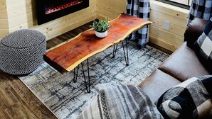 A cozy cabin interior with a wooden interior, a flat-screen TV mounted on a shelf, a fireplace below the TV, a wooden coffee table with a plant, a patterned ottoman, and a plaid-curtained window.