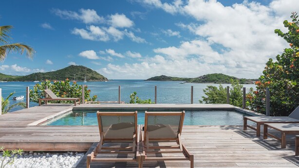 Una terraza de madera con sillones de descanso y una piscina con vista a un mar azul cristalino, donde se ven barcos e islas a lo lejos.