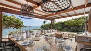 Una mesa de comedor de madera dispuesta con vajilla azul y blanca, rodeada de sillas de mimbre, bajo un techo de madera con luces enredadas colgando, con vistas a una piscina y abundante vegetación.