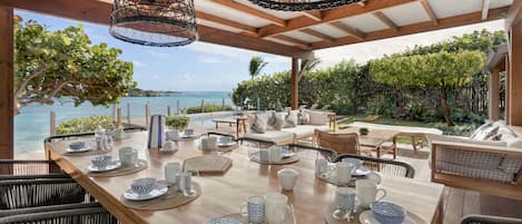 Una mesa de comedor de madera dispuesta con vajilla azul y blanca, rodeada de sillas de mimbre, bajo un techo de madera con luces enredadas colgando, con vistas a una piscina y abundante vegetación.
