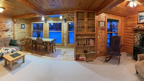 Acogedor interior de una cabaña de madera con comedor, chimenea y estantería.