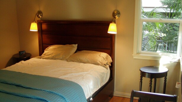 Ein ordentlich bezogenes Bett mit blauer Decke, einem hölzernen Kopfteil, zwei Nachttischlampen, einem kleinen runden Tisch und einem Stuhl.