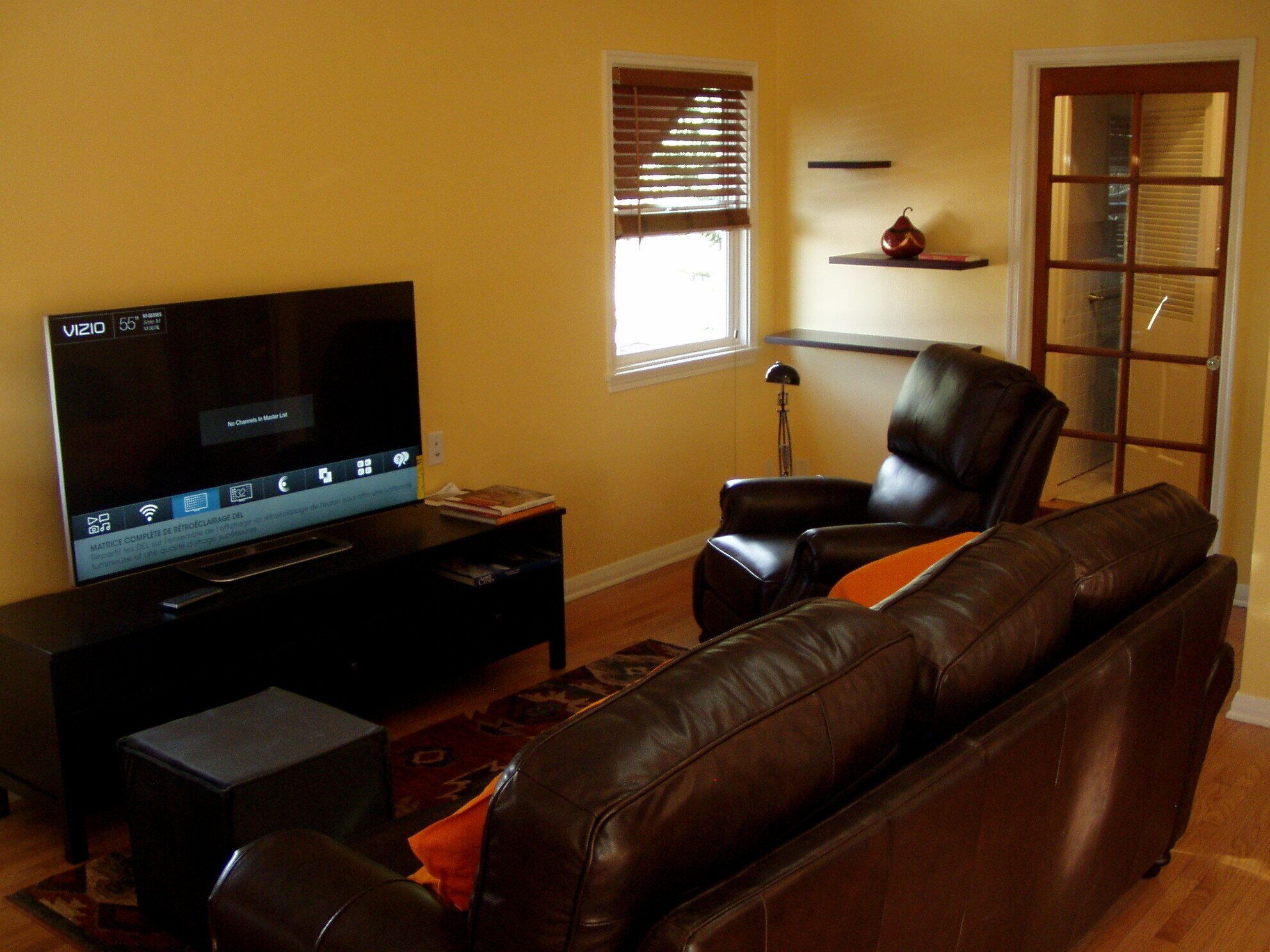 A living room with a flat-screen TV, a leather sofa, a small table, and a window with blinds.
