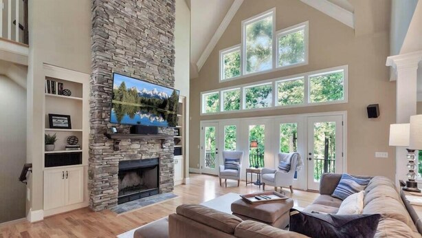 A spacious living room with a stone fireplace, a large flat-screen TV, and a view of greenery through large windows. - Foreststone Fabulous Lake Keowee Waterfront Home (West Union)