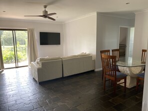Een woonkamer met een bank, een glazen tafel met stoelen, een plafondventilator en een televisie aan de muur.